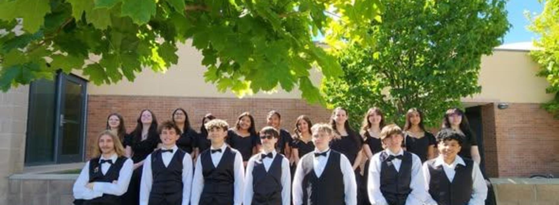 A choir group posed in formal attire outside, with some sitting and others standing.
