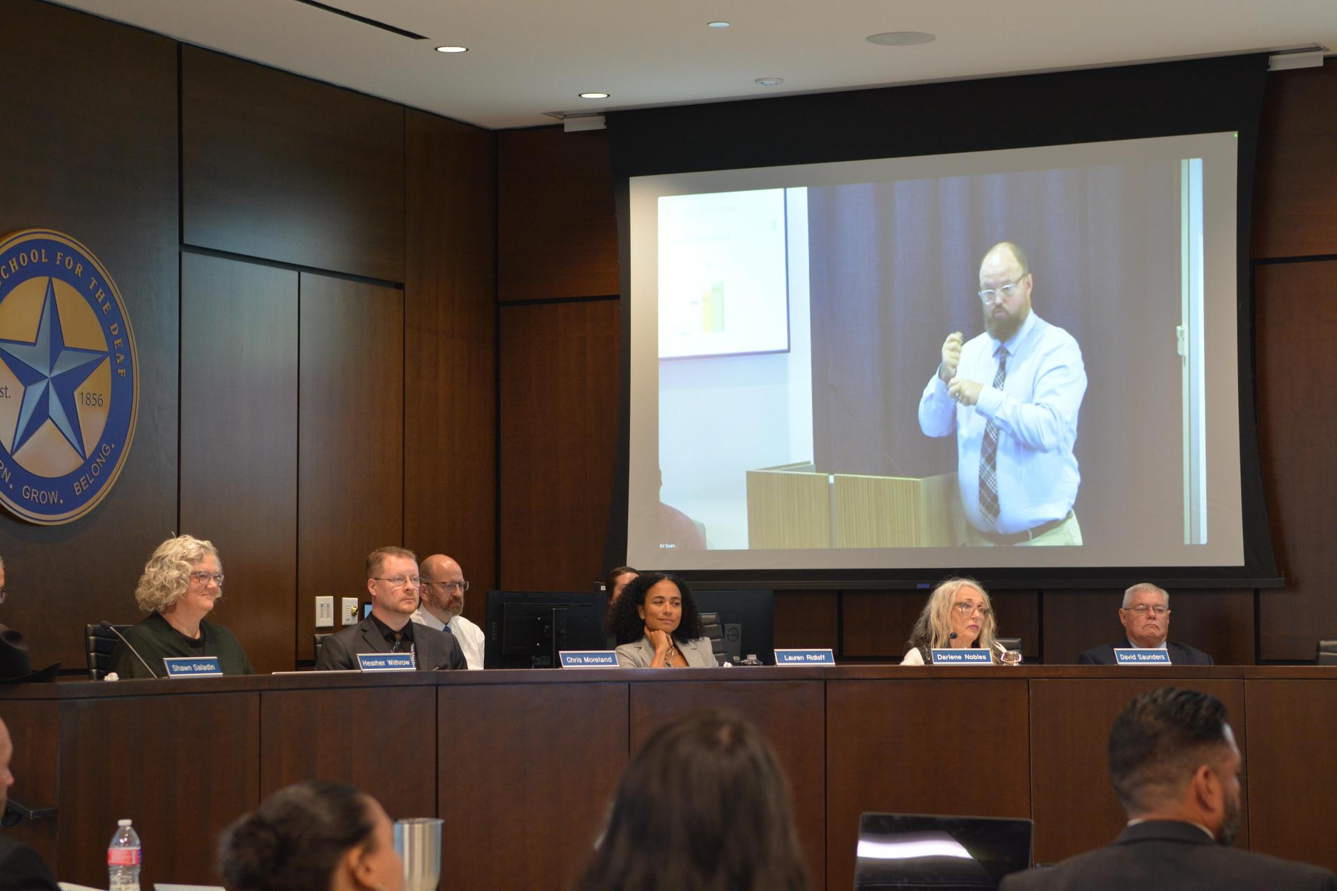 Board members sit at the dais under the Texas School for the Deaf seal listening to presenter.