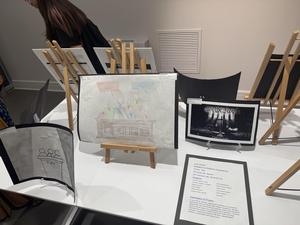 Art pieces on easels showcasing courtroom themes, with a person taking notes in the background.