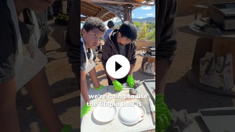 Two students wearing aprons and green gloves working outdoors at a table, preparing tilapia under a wooden canopy.