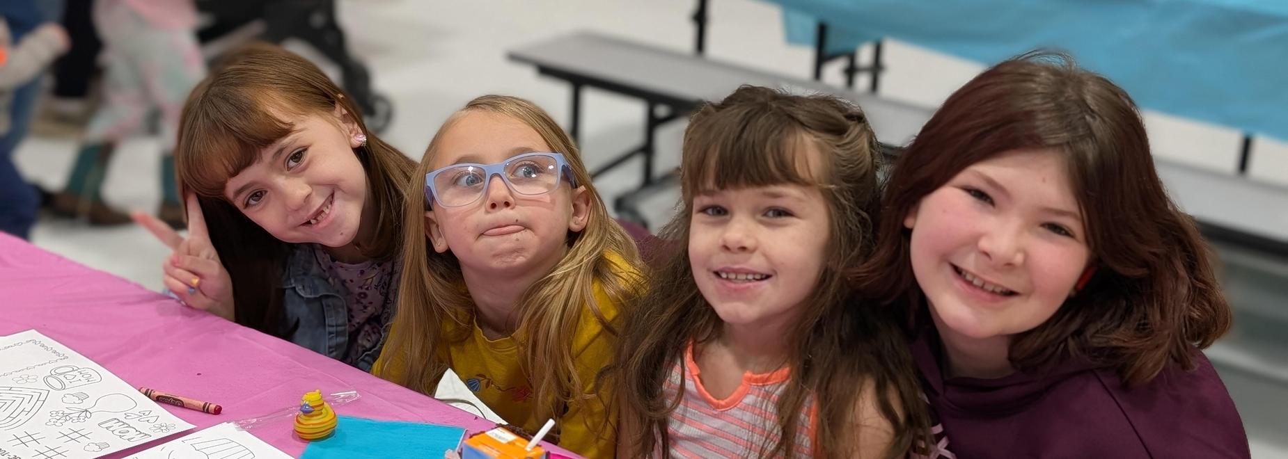 Four girls smiling and posing at a table with craft materials and drawings.