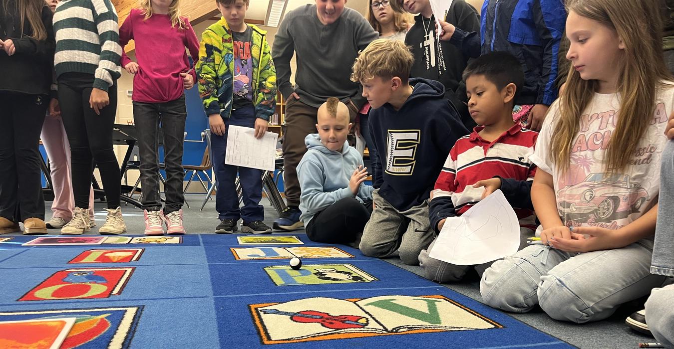 Children gathered around a colorful classroom rug, engaging with a small moving object.