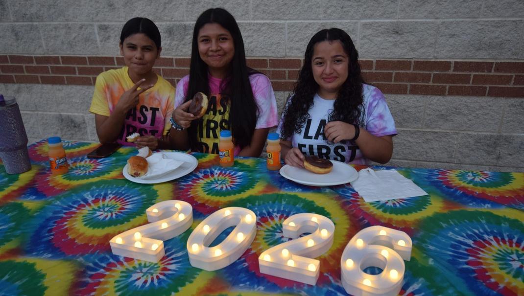 Three girls sitting at a colorful table, enjoying donuts and drinks, with illuminated '2026' sign, celebrating their "Last First Day of School".