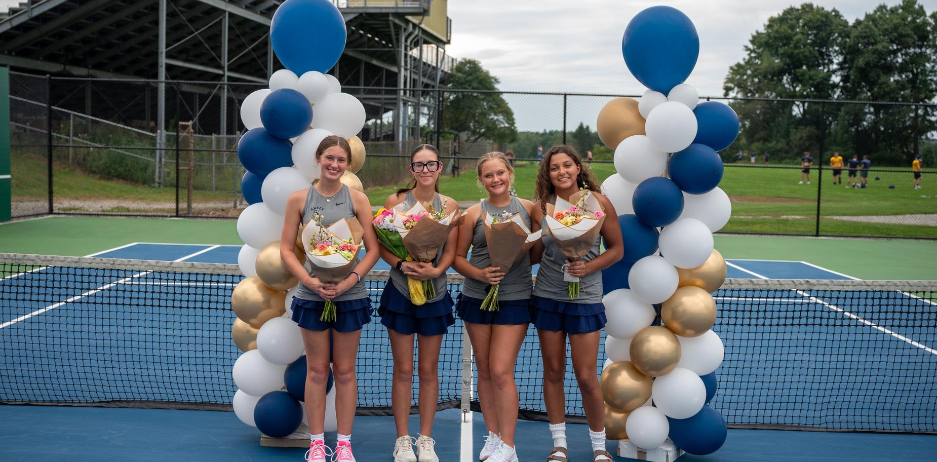 Four girls in tennis outfits holding bouquets near a balloon arch on a tennis court.
