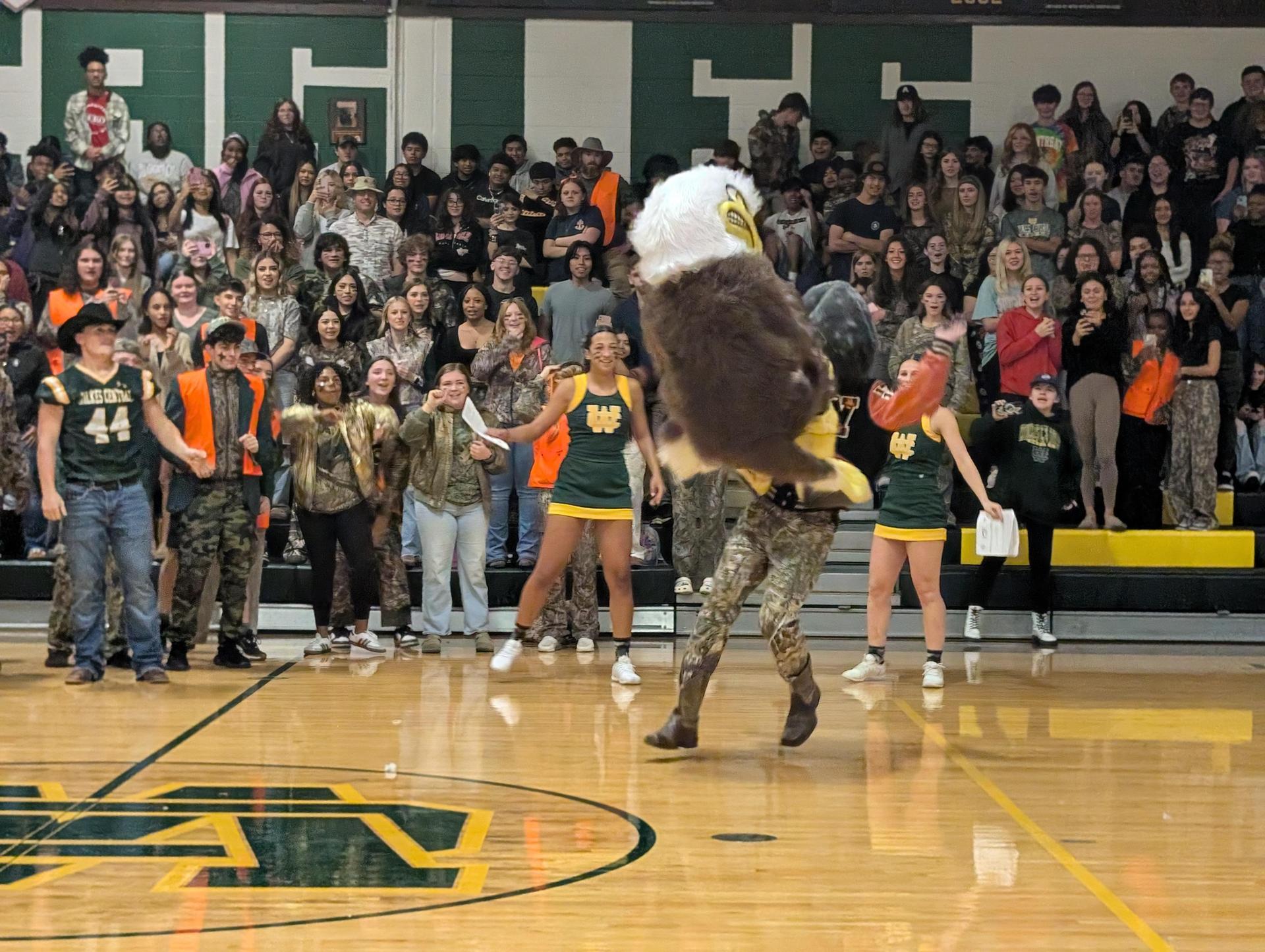 Cheerful crowd celebrates as a mascot performs stunts at a school event.