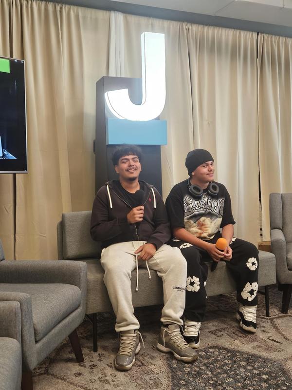 Two young men on a sofa, one holding a microphone and the other holding an orange object.