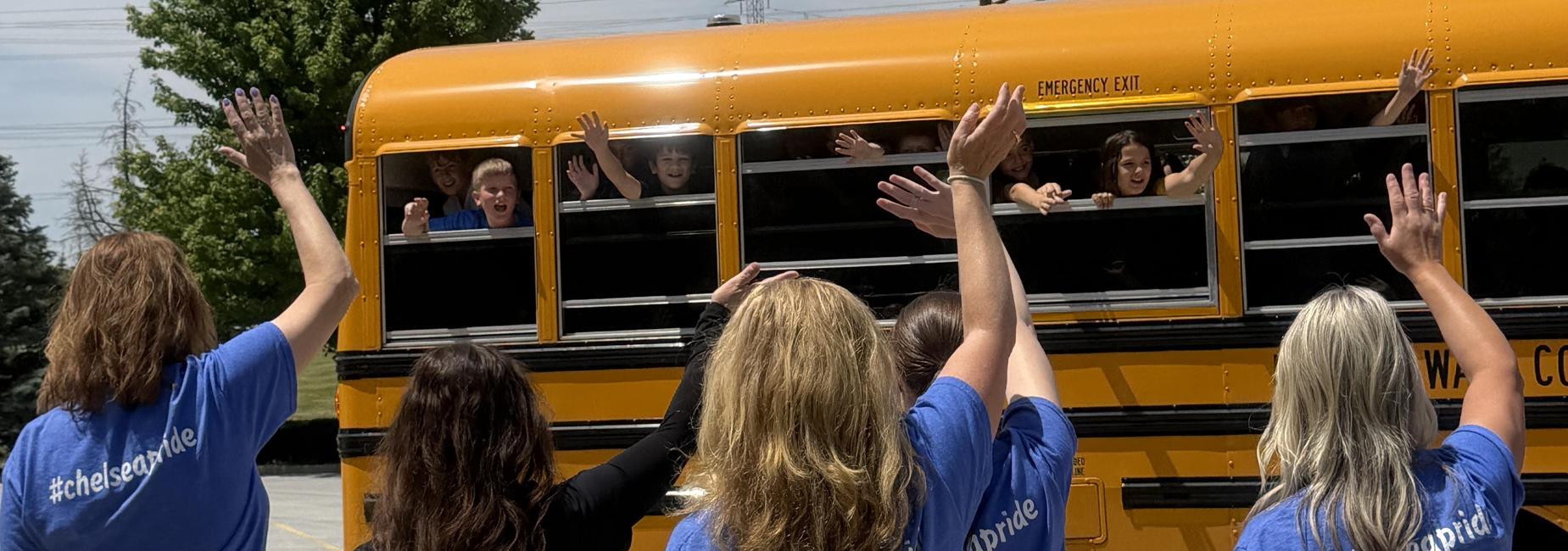 Students and adults wave from a school bus on a sunny day.
