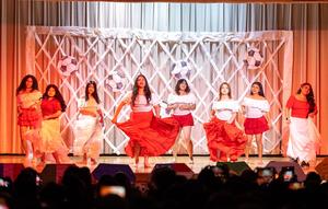 A group of students perform a coordinated dance on a school stage, wearing red and white outfits with flowing skirts. Several dancers lift and swirl their skirts as they move in unison. The backdrop features a white lattice design decorated with soccer ball cutouts, lit by warm stage lighting. Audience silhouettes are visible at the bottom, with some people holding up phones to record the performance.