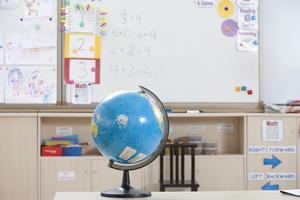 A classroom setting presents an inviting tableau of learning. A blue globe with continents stands prominently on a light table, acting as a visual centerpiece. In the background, a whiteboard displays colorful designs and simple math problems. Labels in clear fonts indicate different learning centers. The overall mood is one of lighthearted engagement, making this image ideal for use in educational materials, school websites, or projects related to geography, education, and early childhood development.