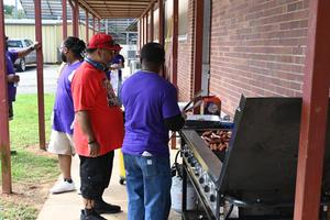 Three men grilling hot dogs under a sheltered area with purple shirts.