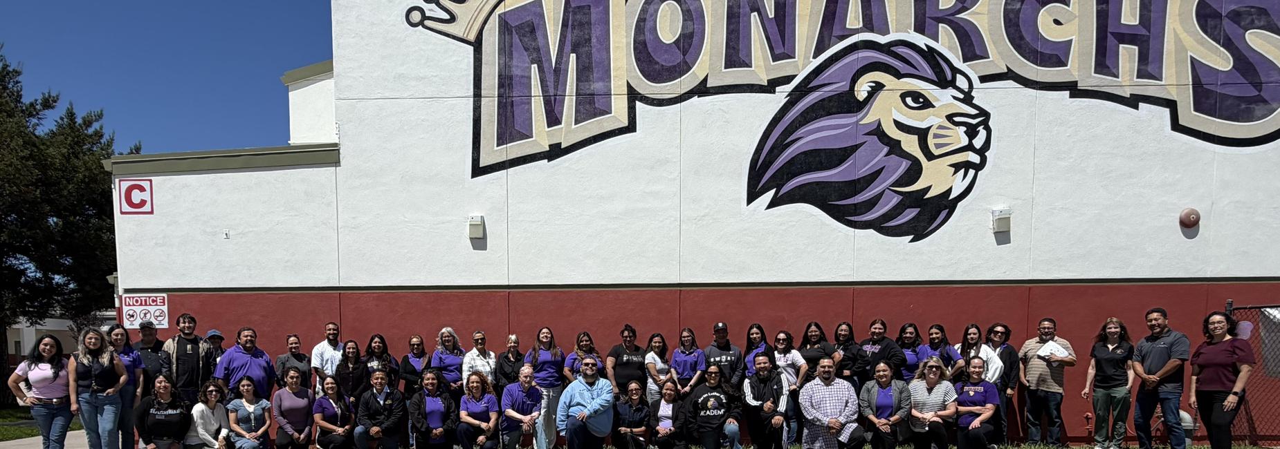 2026-2027 Staff standing in front of "Home of the Monarchs" mural outside cafeteria.