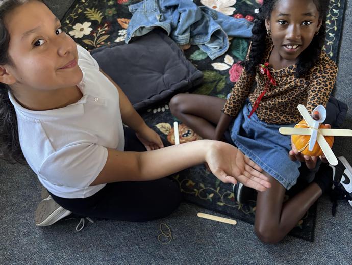 Two girls sitting on a rug, one holding a handmade craft in her hand.