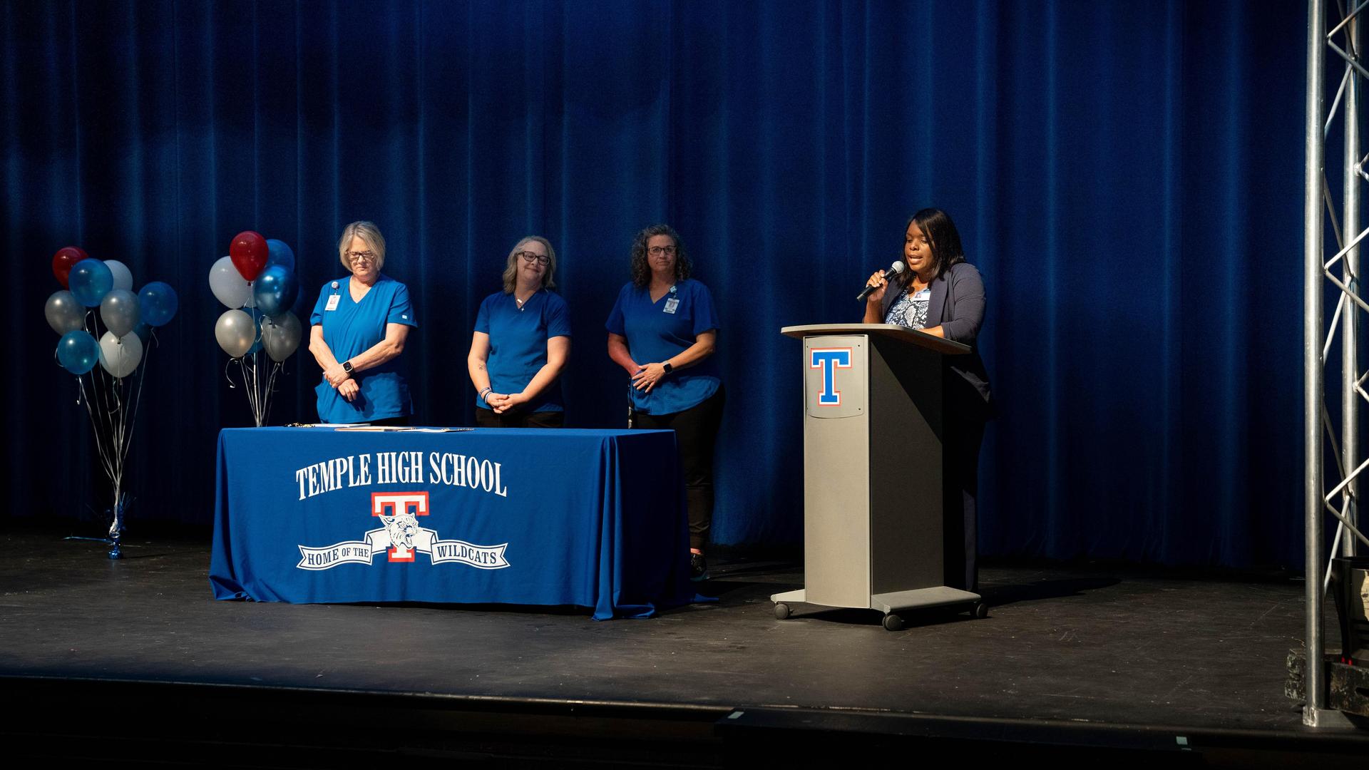 Four people in blue shirts at a podium on stage with balloons and a school banner.