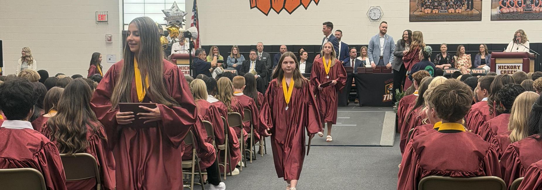 Graduates in maroon gowns walking down an aisle during a ceremony.