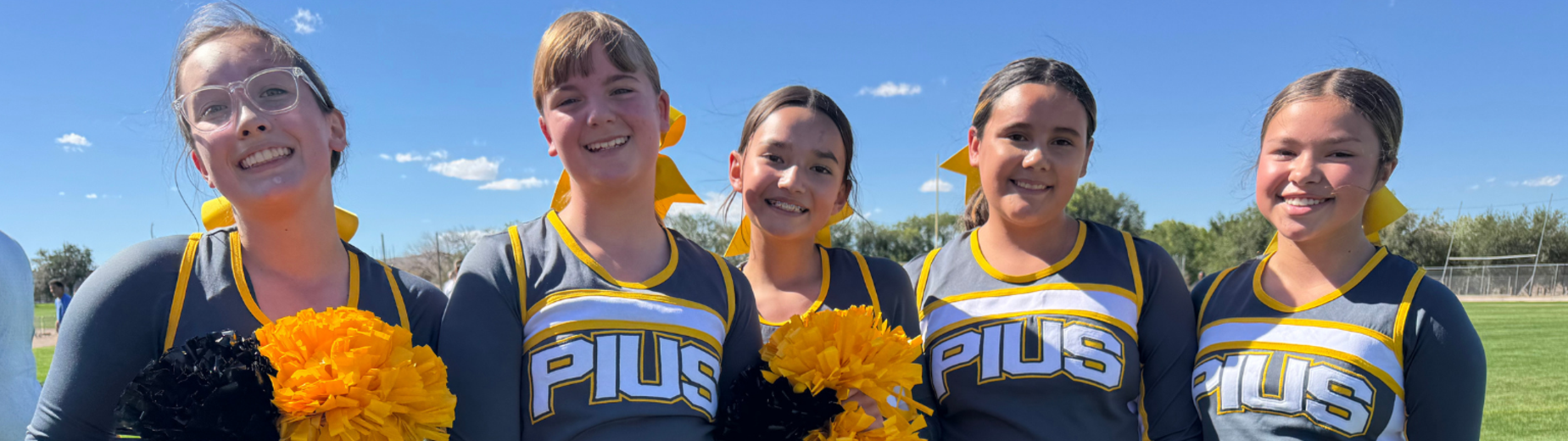 Five cheerleaders in uniforms holding pom-poms on a grassy field under a blue sky.