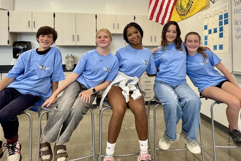 Five students post in blue t-shirts and wave to the camera