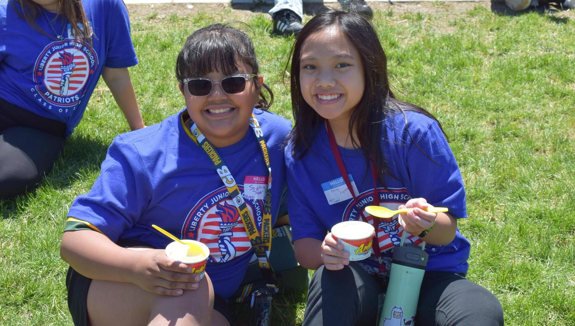 Two smiling girls in blue shirts hold snacks in a grassy area.