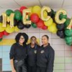 Three individuals pose under a colorful balloon arch spelling 'MECCA'.
