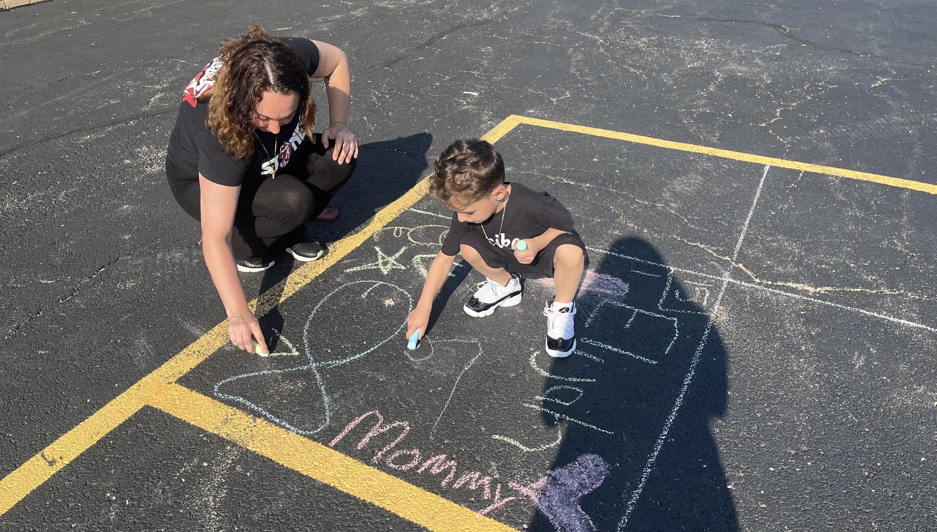 A child and an adult draw with chalk on a parking lot.
