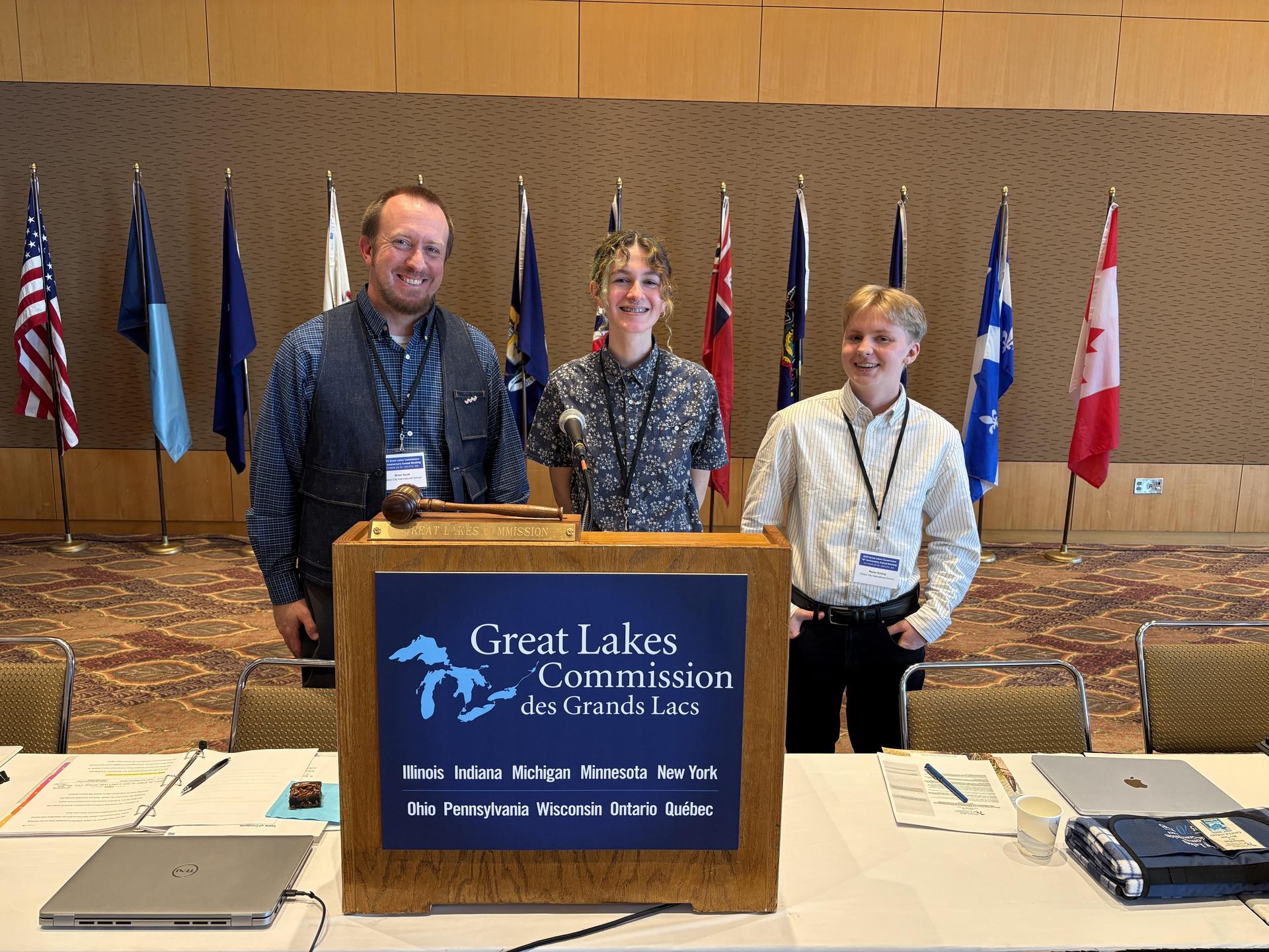 A teacher and two students standing behind a podium that reads "Great Lakes Commission des Grands Lacs"