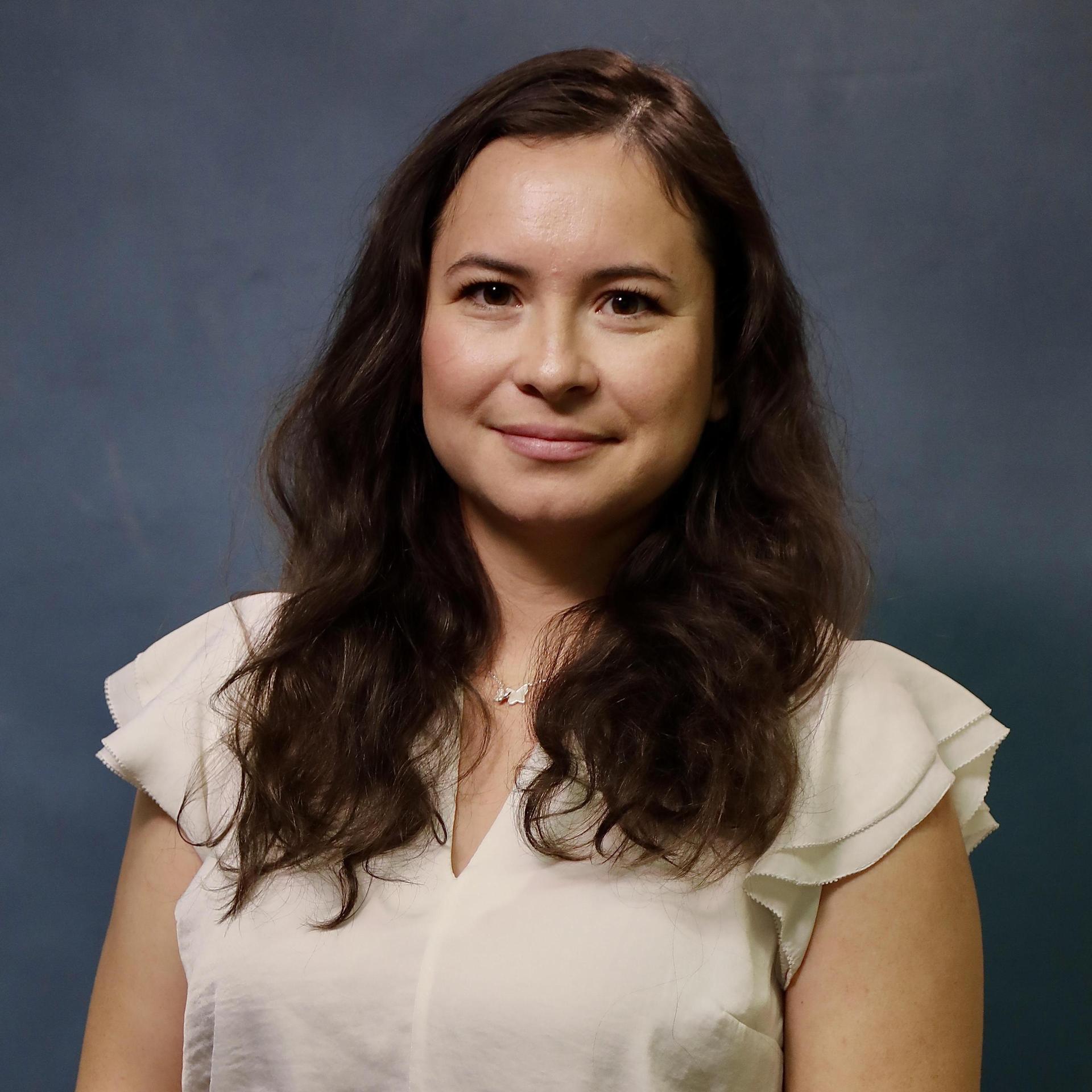 Woman with long, wavy hair wearing a white blouse against a blue background.