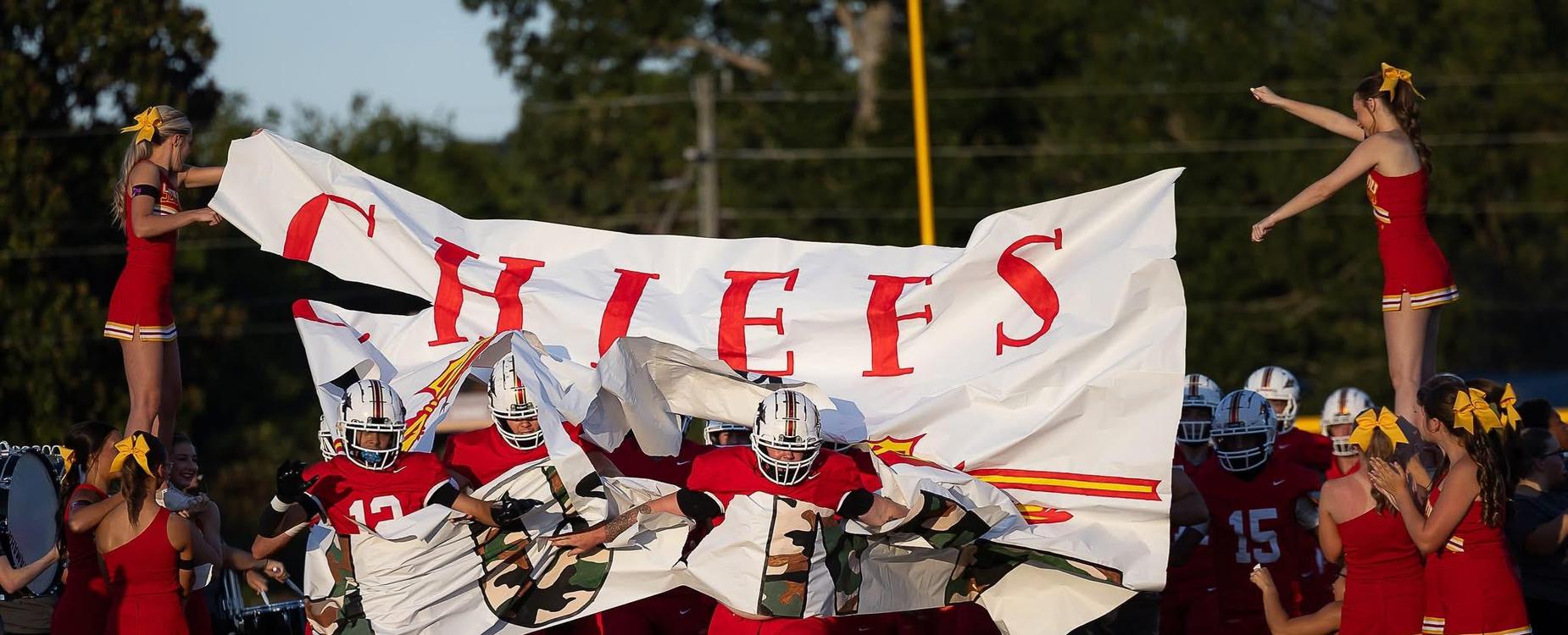 Football players running through a large banner with 'CHIEFS' while cheerleaders cheer.