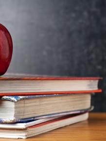 A red apple atop a stack of three books on a wooden surface.