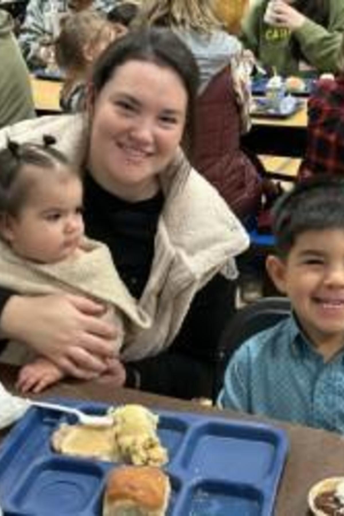 A woman holds a baby while sitting at a table with a smiling child and food trays.