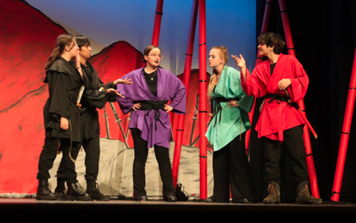 Students performing in the Texas School for the Deaf (TSD) senior theater production of The Seventh Blade, an original story by TSD student Peyton John Yevich. The actors, dressed in vibrant martial arts-inspired costumes in shades of black, purple, green, and red, are engaged in an intense scene. The backdrop features red mountains and tall bamboo stalks, evoking a dramatic and immersive setting.