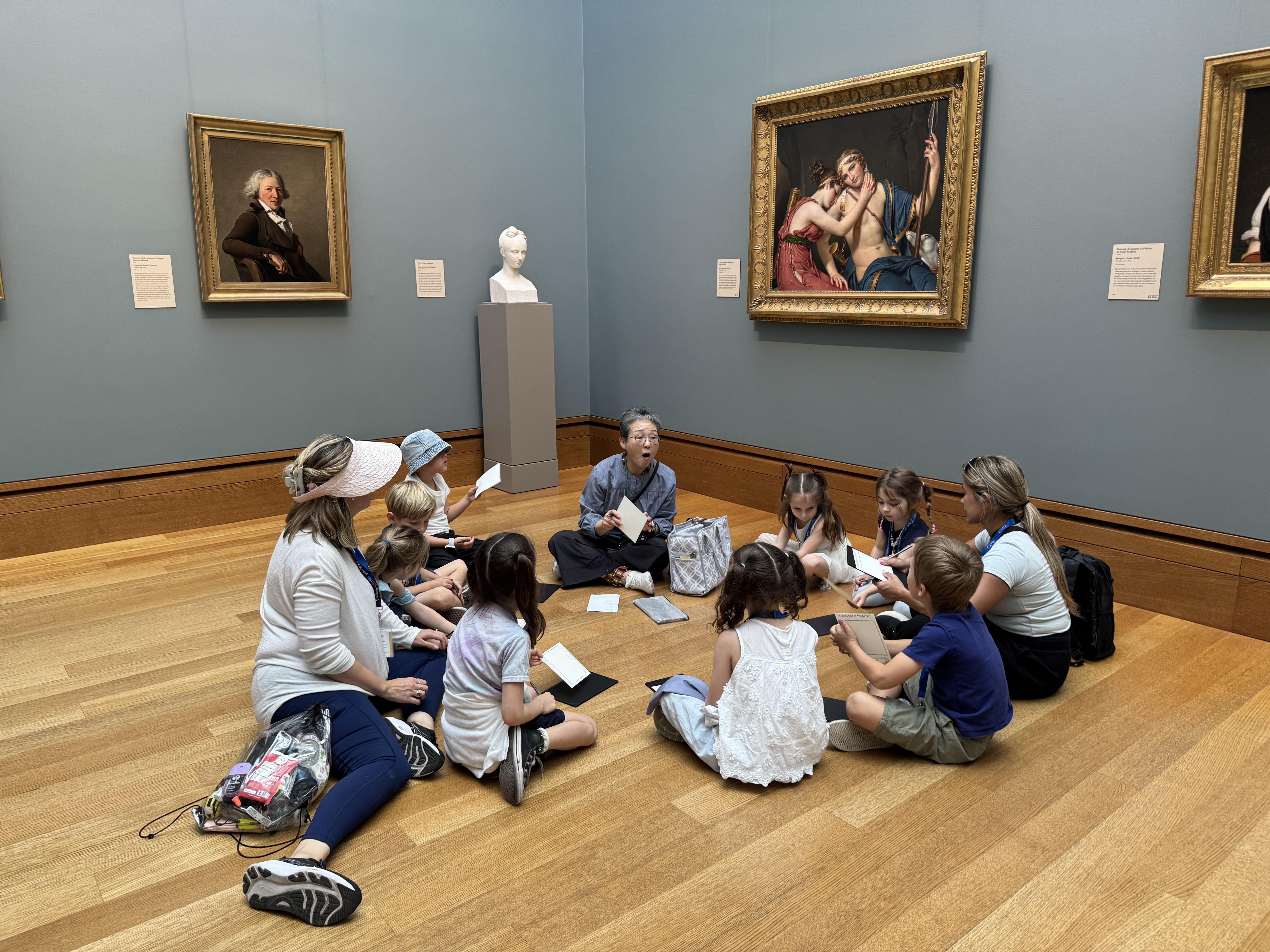 kindergarten students sitting on the floor of a museum with 3 adults listening to the docent talk about the art.
