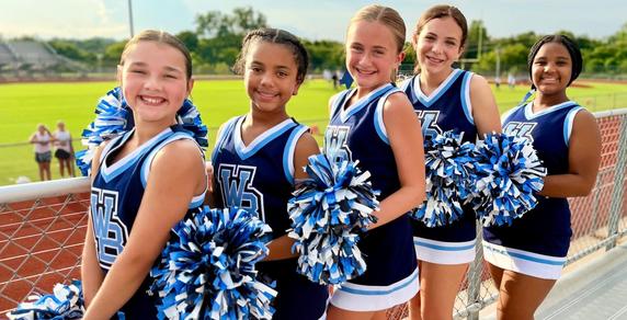Five cheerleaders pose happily with pom-poms in a sports setting.