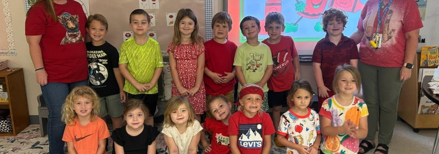 Group of children and two adults in a classroom, all wearing red clothing.