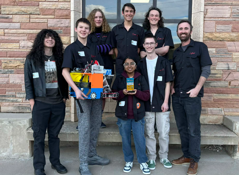 A group of high school students and their teacher pose for a photograph. One student is holding an award, and one is holding a robot.
