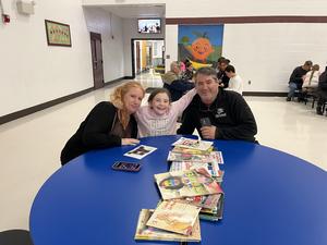Two adults and a child smiling at a blue table filled with books in a school setting.