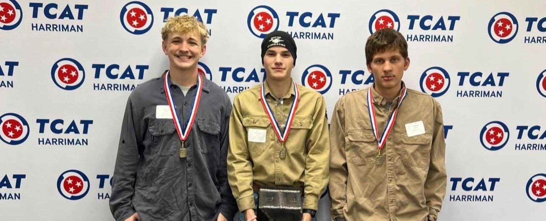 Three students proudly displaying medals and holding a trophy at a TCAT event.