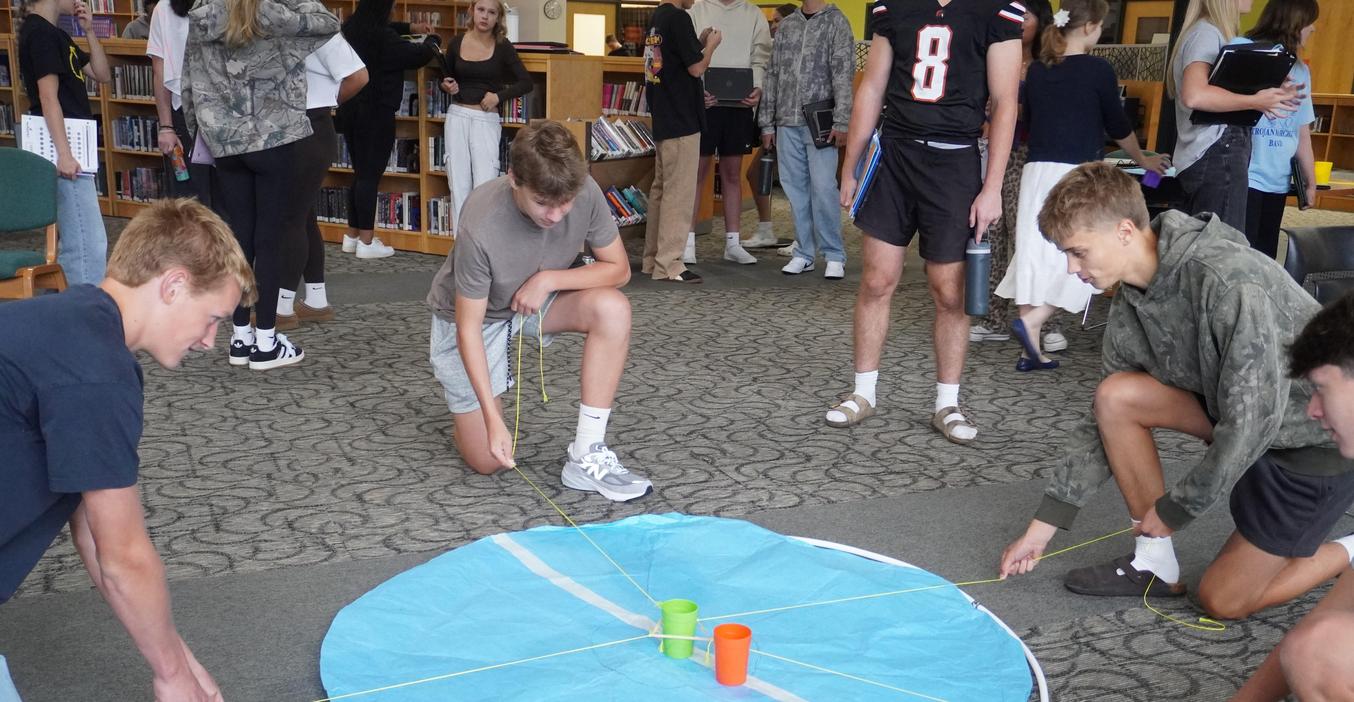 Four high school students work together using rubber bands to solve a physics challenge moving a cup from outside a circle.