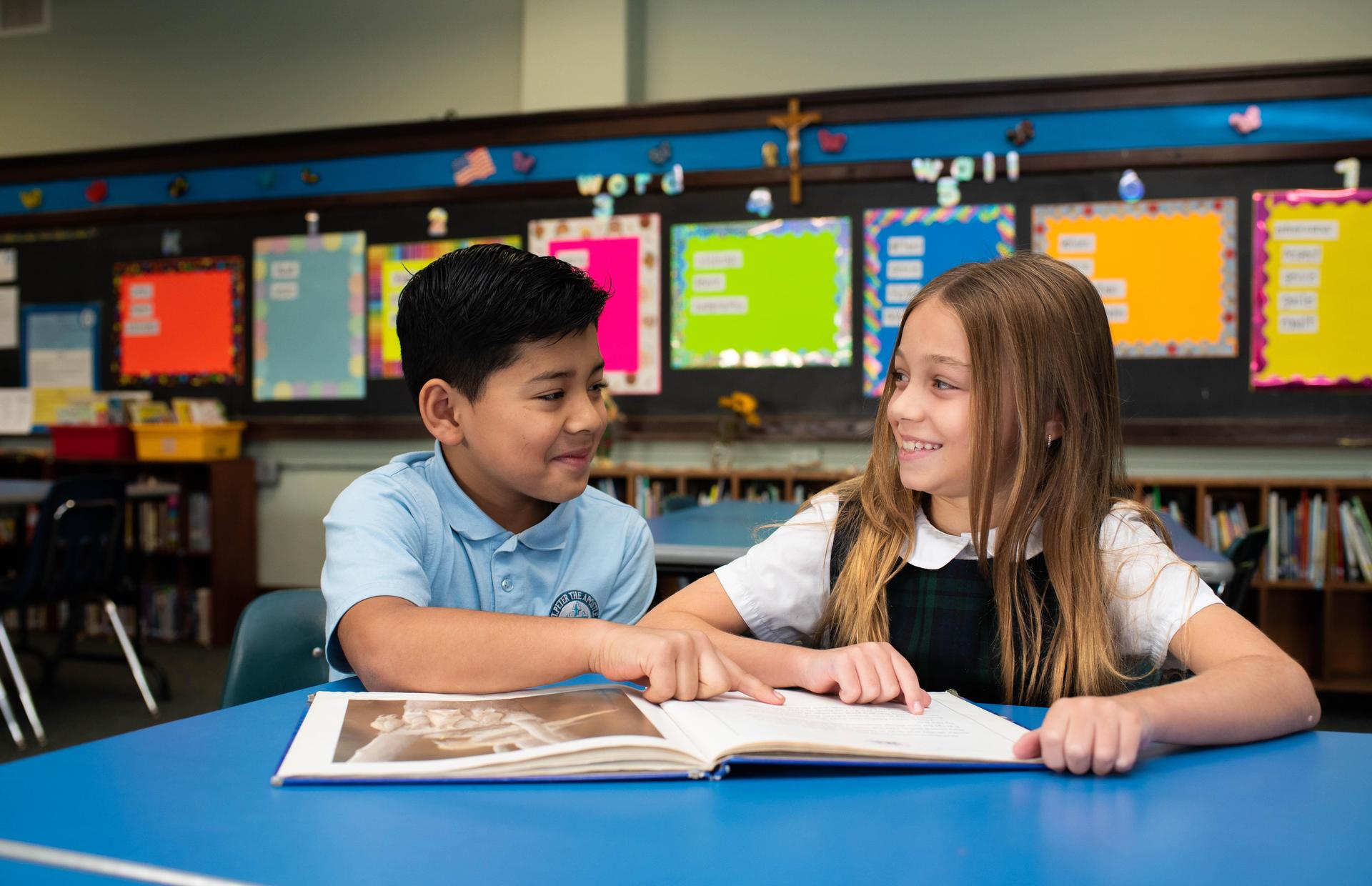 Two children share a book at a table in a classroom, smiling and engaged.