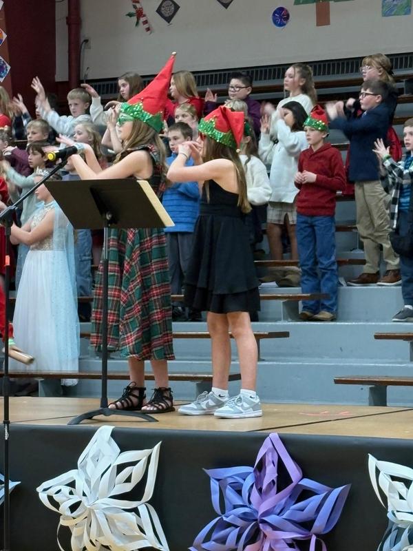 Two girls sing on stage in festive holiday dresses with students cheering behind them.