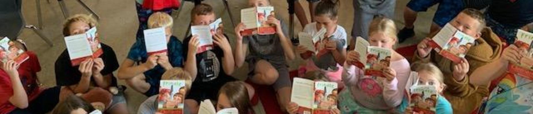 Group of students sitting on floor of classroom with books in front of their faces