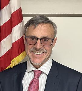 Image of Marc Cohen wearing a suit and tie standing next to the US flag