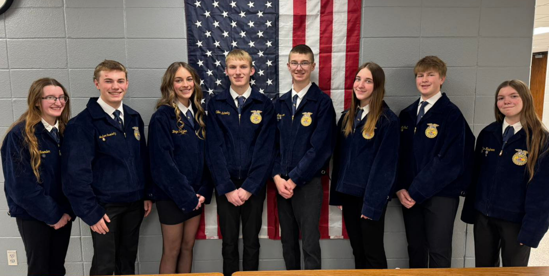 Group of students in blue jackets standing in front of an American flag.