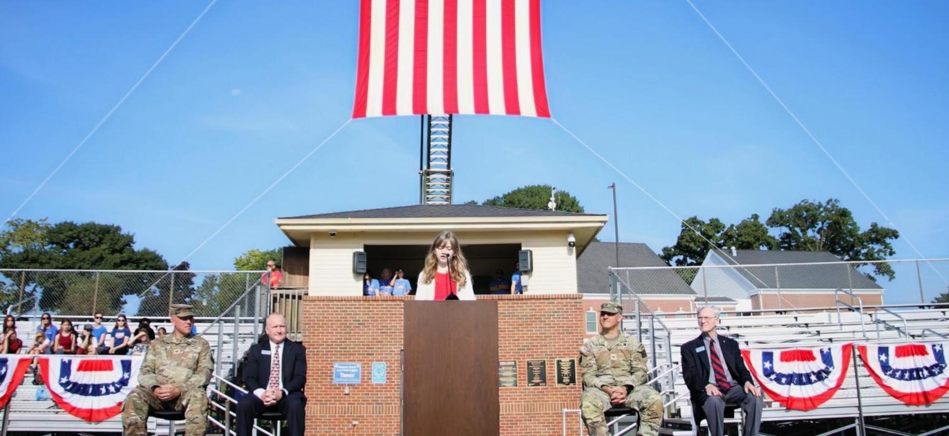 Girl speaks at a podium under a large American flag with service members present.