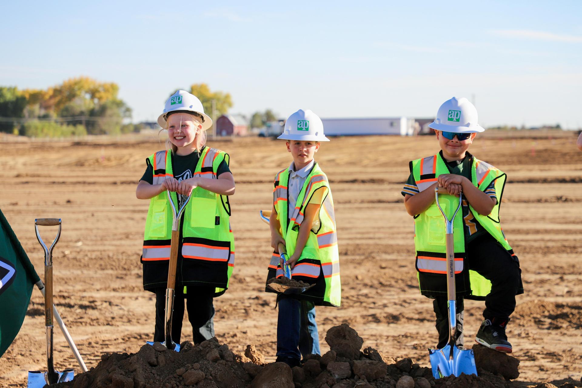 Three children in safety gear posing with shovels beside a mound of dirt.