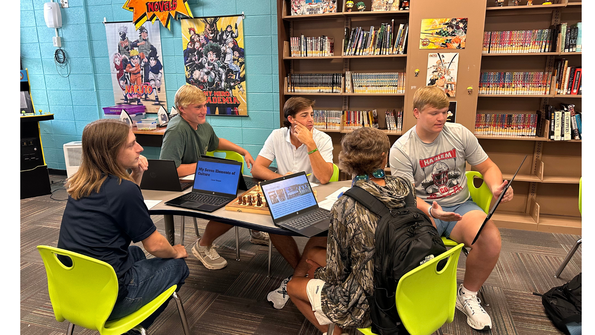 Group of students gathered around a table playing chess and discussing in a library.
