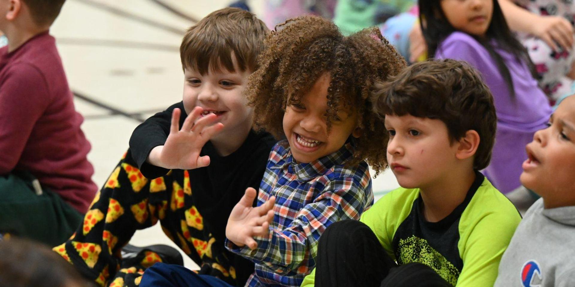 Three children sitting together, smiling and waving during an event.