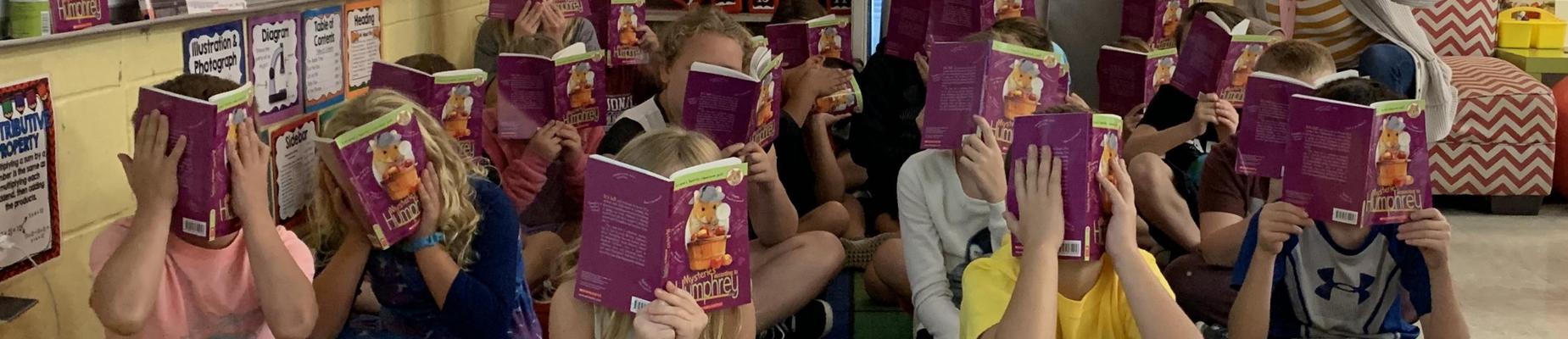 Students sitting on floor in classroom with books in front of their faces