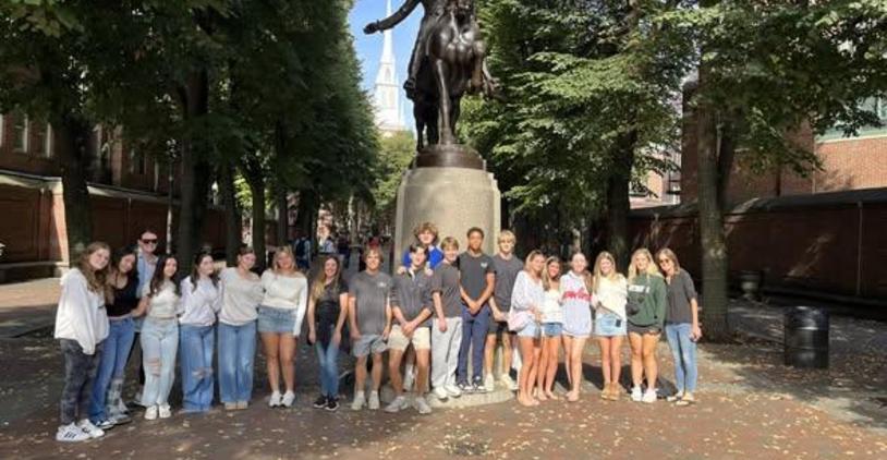 A diverse group of people poses for a photo in front of a statue on a tree-lined street.
