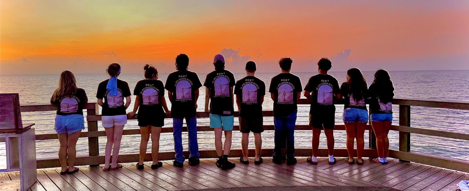 Group of eight people in matching shirts watching a sunset by the ocean.