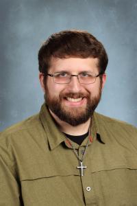 Smiling man in a green shirt with glasses and a cross necklace.
