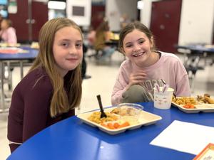 Two smiling girls sit at a table with trays of food in a cafeteria setting.